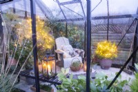 Recycled plastic chair with blankets and cushions, wooden crate and plants adorned with fairy lights inside a greenhouse