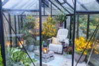 Recycled plastic chair with blankets and cushions, wooden crate and plants adorned with fairy lights inside a greenhouse