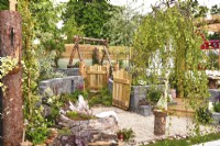 View of the woodland inspired garden with open wooden gates leading to different area, large logs, a decorative Connemara walling system, swing. Planted with  Astilbe koreana, Trachelospermum jasminoides, Heuchera 'Licorice', Salix elaeagnos, Betula pendula. June
Designer: Mary Anne Farenden. Bord Bia Bloom, Super Garden, Dublin, Ireland.