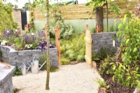 View of a open gate leads into a grass area for dogs with raised bed  made of Connemara wall system and gravel surfaces  in woodland inspired garden surrounded by a wooden planks fence. June

 


