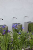 Three chrome water spouts cascading into metal containers, bordered by purple iris and ferns in the 'Sociability' garden at BBC Gardener's World Live 2015, June