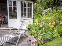 Country garden summerhouse and patio with lounger and terracotta succlent pots next to a mixed border featuring Tulipa 'Sunny Prince' and spring foliage