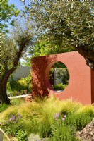 A orange compartment wall with a circular open window, underplanting with Stipa tenuissima and  large trees of Olive in Mediterranean garden. June
Designer - Alan Rudden
