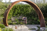 Metal table and chairs on paving under a Corten steel arch  - 'It Doesn't Have to Cost the Earth' - designer Michael Lote - RHS Malvern Spring Festival 2024