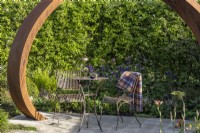 A Corten steel helix sculpture curves over a metal table and chairs with perennial border and Crataegus monogyna hedge behind on 'It Doesn't Have to Cost the Earth' - designed Michael Lote - RHS Malvern Spring Festival 2024